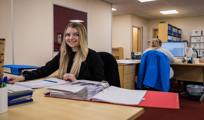 A business apprentice at their desk with papers and files on whilst looking towards the camera