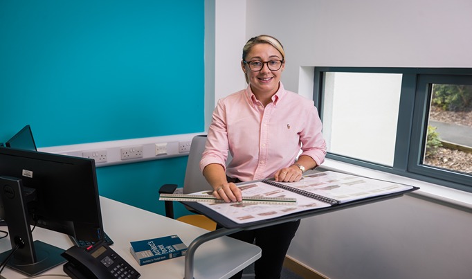 A Construction Apprentice in their office looking towards the camera with plans on the stand up table below her