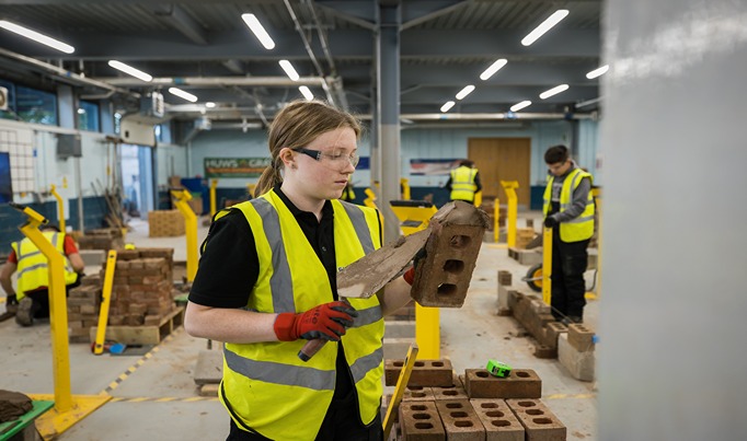 A brickwork student at Bersham road in PPE applying cement to a brick ready to lay it down