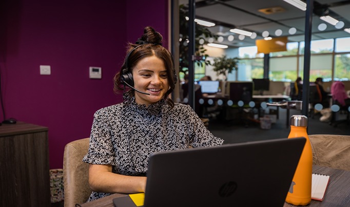 A customer service apprentice for Moneypenny sat at a desk on her laptop with a headset on