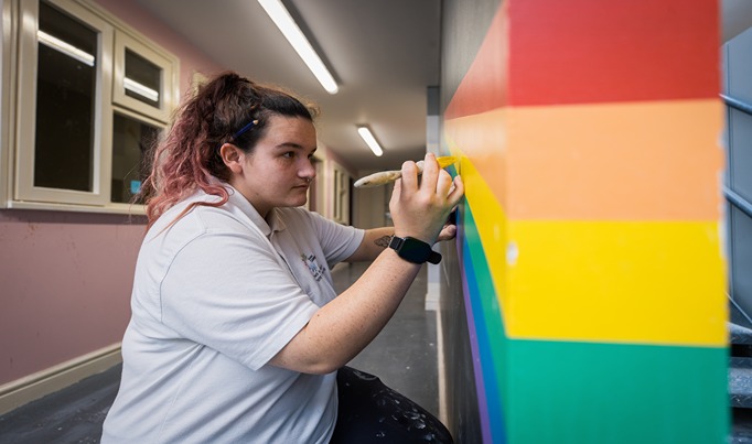 A painting and decorating student painting yellow onto a rainbow coloured wall