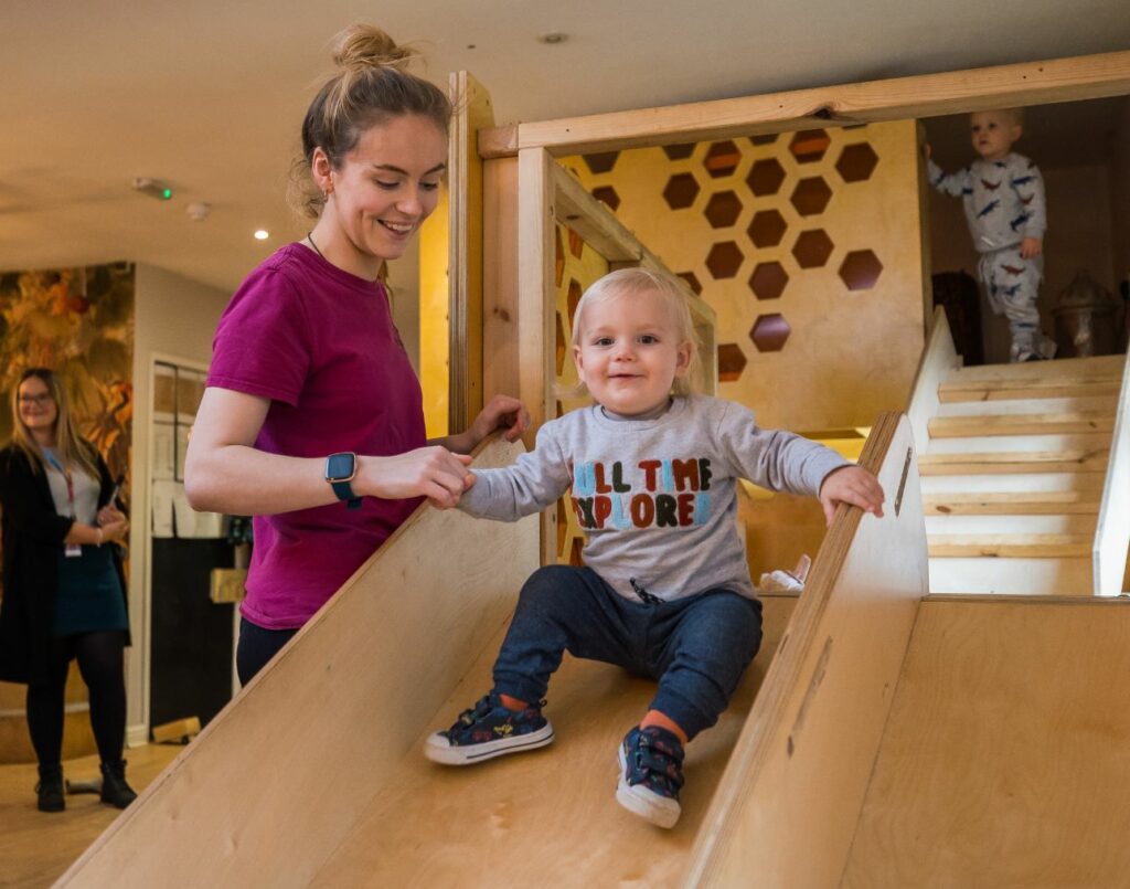 A childcare apprentice at The Homestead helping a child down a wooden slide