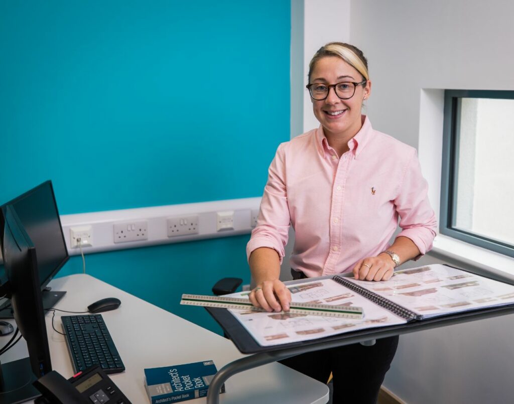 A Construction Apprentice standing in their office looking towards the camera with plans on the stand up table below her