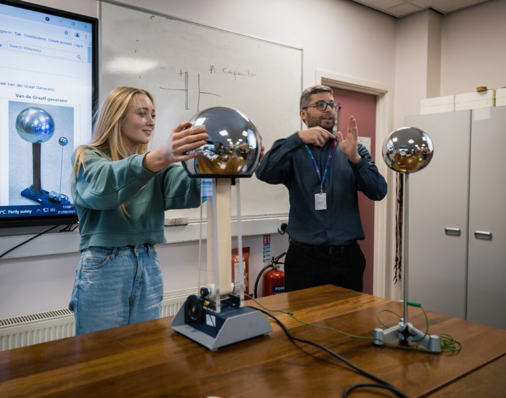 A student at the front of a class with their hands on a Van De Graaff generator, stood by the tutor