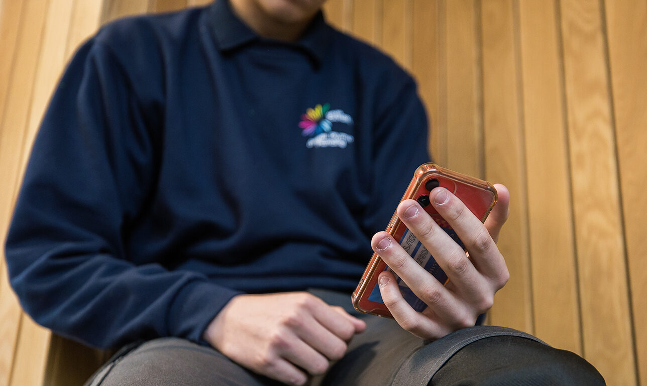 A student sitting down holding their phone wearing a Coleg Cambria jumper