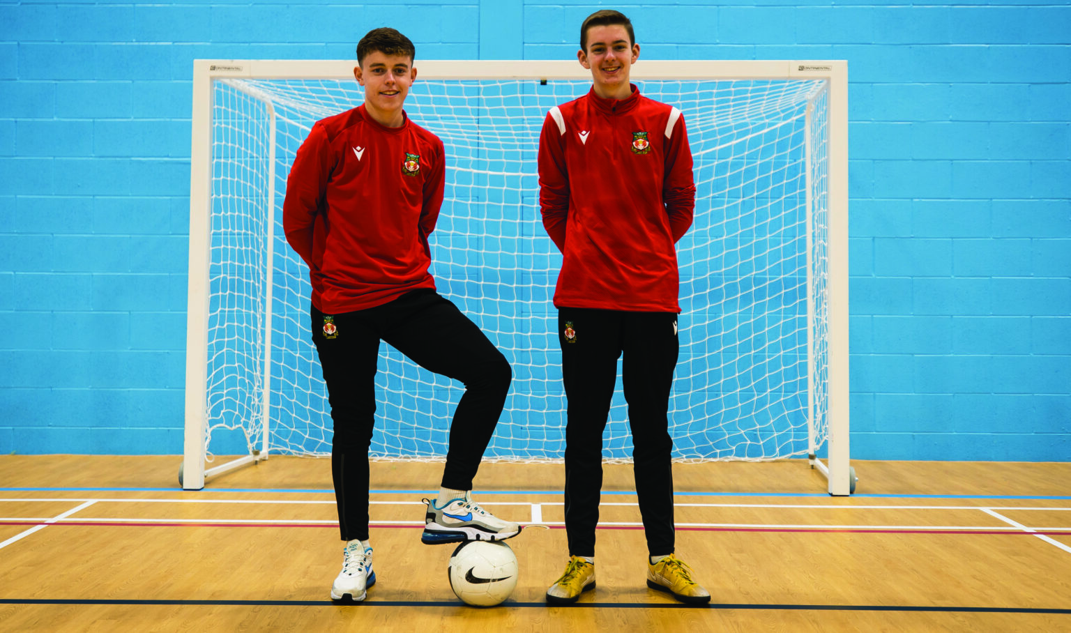 Two students standing in front of an indoor football goal in Wrexham football tracksuits