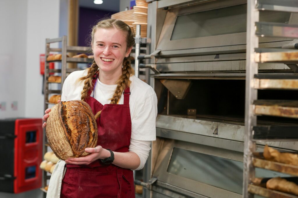 ial Bakery apprentice Ella Muddiman taking a hero shot holding a loaf of sourdough