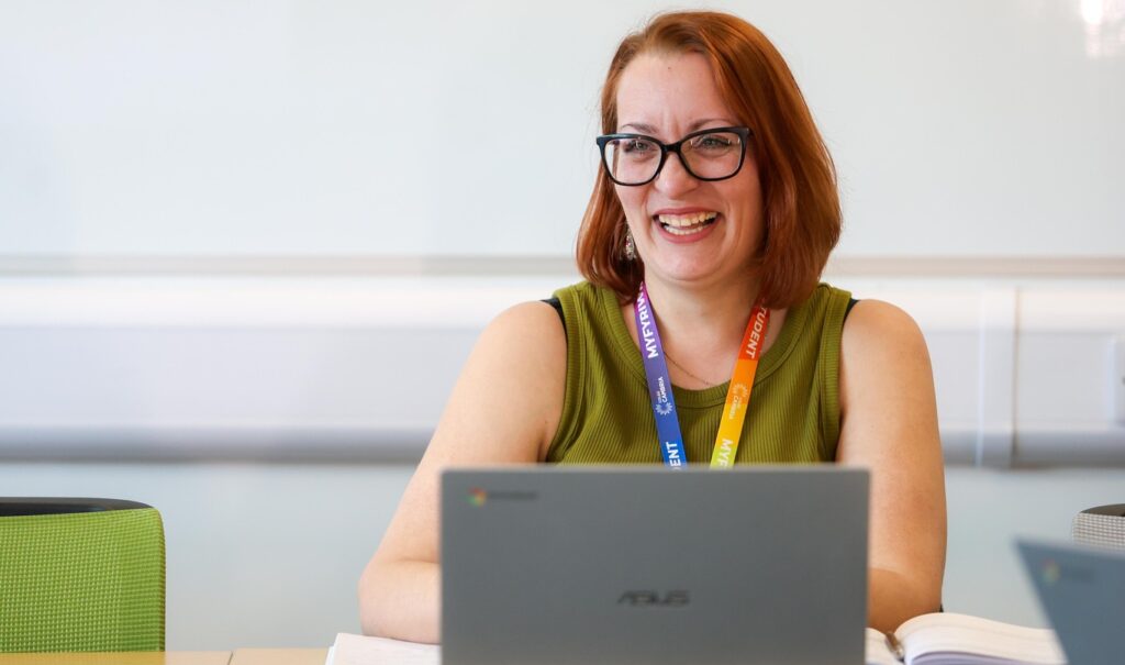 A close up of an adult learner sat at a desk with a big smile with a laptop in front of them