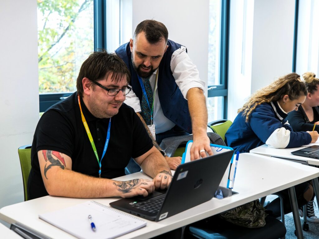 an adult tutor standing behind an adult learner who is sat down on their laptop at a desk which the tutor is pointing too