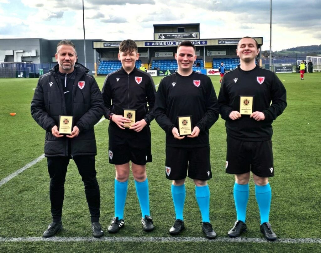 Bradley Tonks (fourth official) with Cambria students Dyfan Roberts, Jamie Monks and Levi Roberts in officiating kit standing for a photo for the news post 'A Proud Moment as Three Learners Kick Off Their Officiating Careers at International Youth Match'
