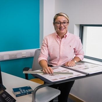 A Construction Apprentice in their office looking towards the camera with plans on the stand up table below her