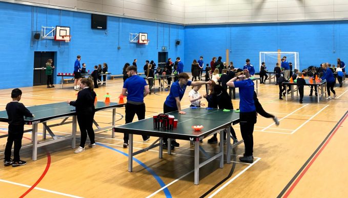 Students and young kids in a sports hall playing table tennis, the image is for the news post 'Coleg Cambria rallied behind a popular tennis academy which went from strength to strength in 2024'